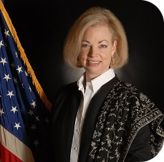 A woman with blond hair wearing a black and white outfit stands in front of a U.S. flag, facing the camera and smiling.