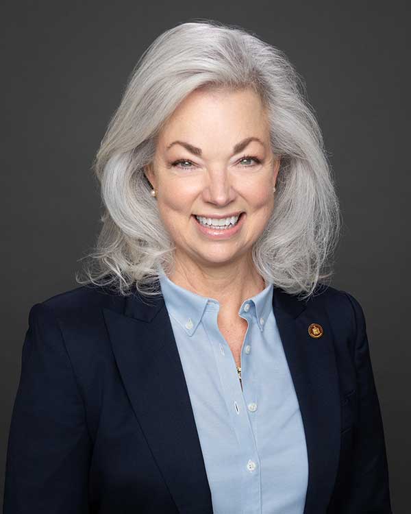 A woman with grey hair smiles while wearing a navy blazer and light blue shirt against a dark background.