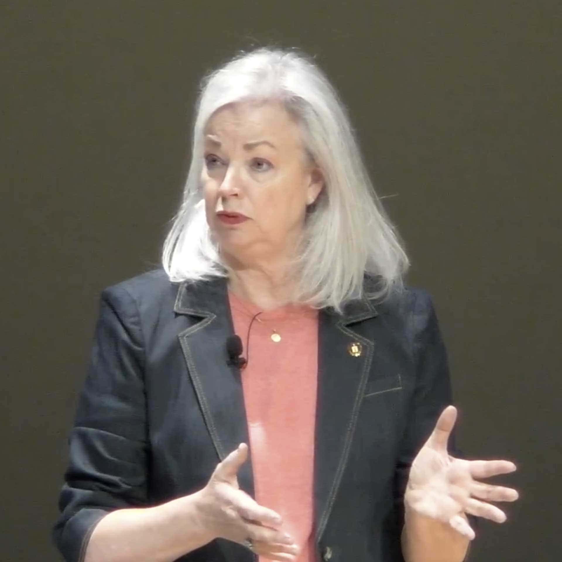 A woman with gray hair speaks while gesturing with her hands. She wears a dark blazer over a pink top and stands against a plain background.