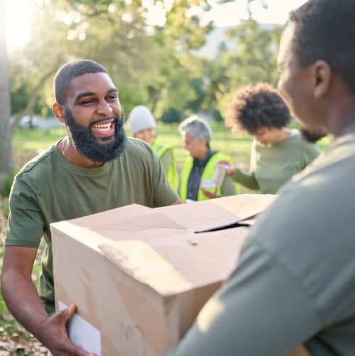Two people smile while carrying a cardboard box outdoors. Others in the background wear safety vests and help, suggesting a community volunteer event.