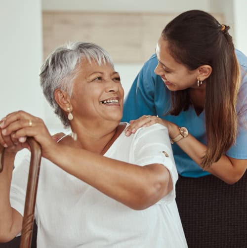 An older woman with gray hair smiles while holding a cane, as a younger woman stands beside her, smiling and touching her shoulder.