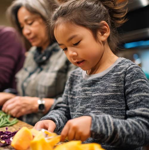A young child cuts yellow vegetables at a table while an older woman prepares food beside her in a kitchen setting.