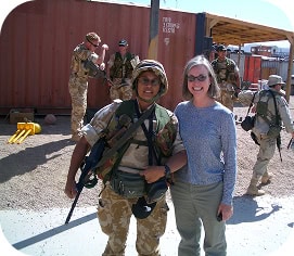 A soldier in uniform stands next to a smiling woman in casual clothes with other soldiers and equipment visible in the background.